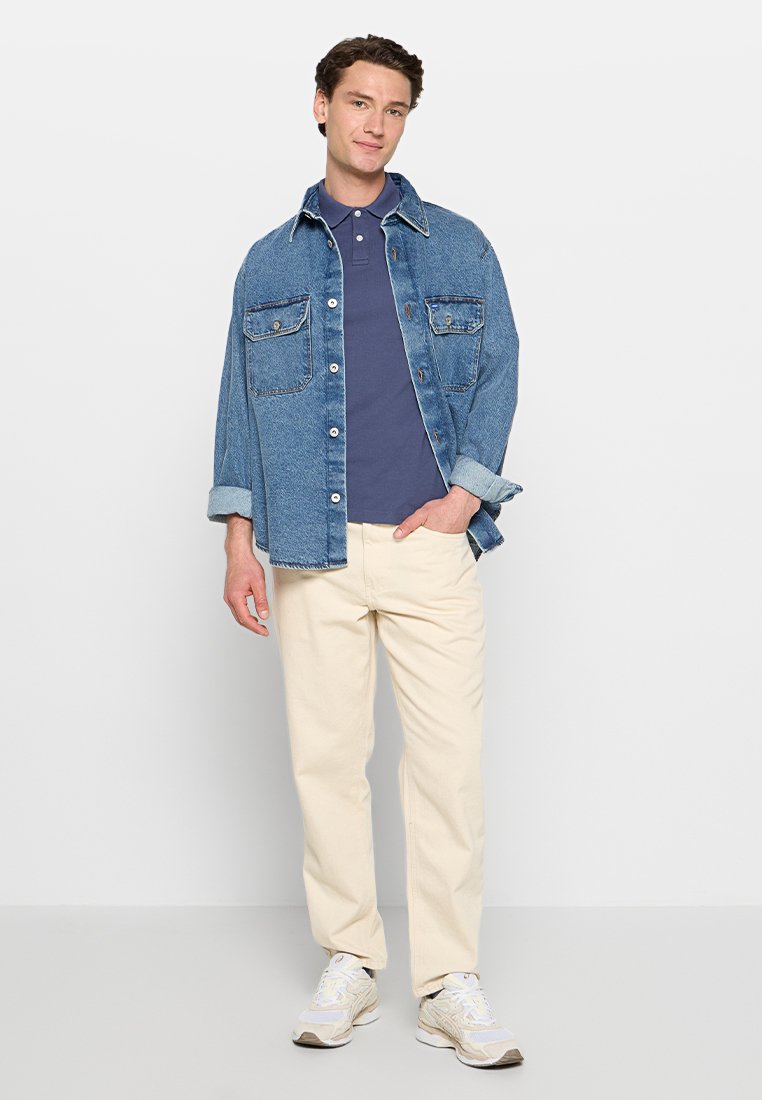 Chemise en denim avec deux poches poitrine, superposée à un polo bleu marine, associée à un pantalon crème et des baskets claires.