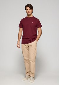 Bordeaux T-shirt met korte mouwen en een klein logo, gecombineerd met beige broeken en lichte sneakers, tegen een neutrale achtergrond.