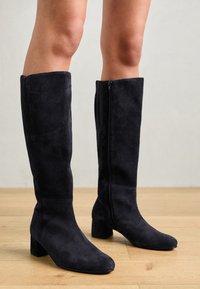 Botas de ante negras hasta la rodilla con punta cuadrada y tacón grueso. Cuentan con una cremallera lateral y textura suave. Fondo con madera clara.