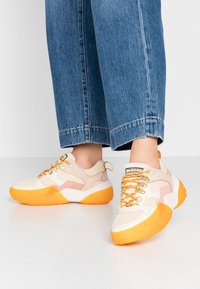 Beige Sport Schuhe mit orangefarbenen Sohlen, strukturiertem Mesh-Obermaterial und metallischen Beschlägen. Schnürdesign mit Akzentpaneelen in Hellrosa.