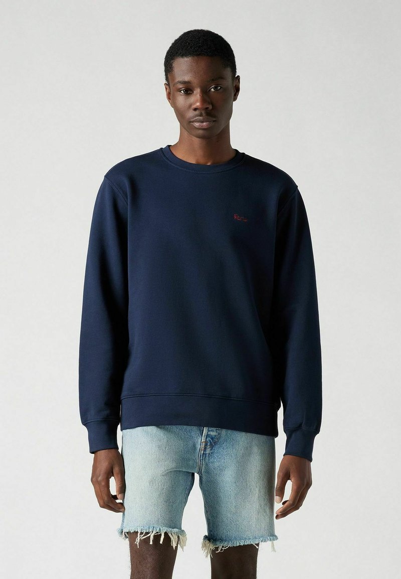 Marineblauwe katoenen sweatshirt met lange mouwen en geribbelde boorden, voorzien van een klein rood logo op de borst; gecombineerd met versleten denimshorts.