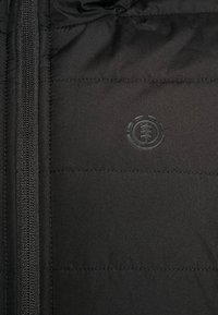 Sort polstret jakke med lynlås og et cirkulært logo i tone-i-tone design. Glat tekstur med horisontale sømdetaljer.