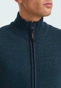 Mørke teal genser med høy ribbestrikket krage, halv-zip lukking og strukturert strikkemønster. Har en svart glidelås med en logotag.
