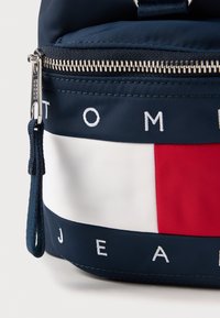 Ναυτικό σακίδιο από νάυλον με ασημένιο φερμουάρ, που διαθέτει οριζόντια κόκκινη και λευκή ρίγα, και το λογότυπο "TOMMY JEANS" κεντημένο σε λευκό.