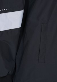 Black jacket featuring a horizontal silver reflective strip and subtle dotted accents, with a front pocket and textured fabric.