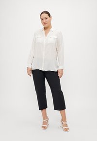 Chemise blanche à manches longues avec des rayures verticales, deux poches poitrine ; assortie à un pantalon noir cropped et des escarpins blancs à lanières.