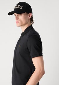 Black polo shirt with a textured collar and a small emblem. Paired with a black cap featuring light gray branding, shown in profile.
