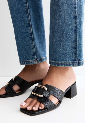 Voeten met zwarte sandalen met een gestructureerde blokhak en gouden gespsieringen, gecombineerd met cropped blauwe spijkerbroeken op een witte ondergrond.