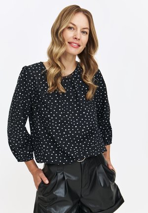 Czarna bluzka w białe kropki, okrągły dekolt, detal z marszczeniem i długie rękawy. Połączona z wysokimi, czarnymi szortami z syntetycznej skóry.