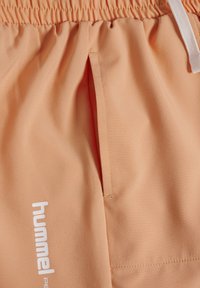Pfirsichfarbene Sportshorts mit elastischem Bund, einer Vordertasche und einem weißen Logo. Hergestellt aus einem glatten, leichten Material.
