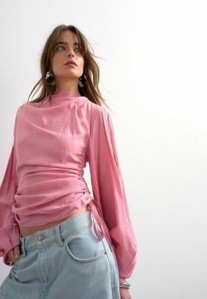 Vrouw draagt een geplooide roze blouse met lange mouwen en lichtblauwe jeans, kijkt iets omhoog tegen een effen witte achtergrond.