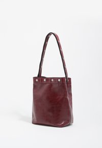 Bordowa skórzana torba typu tote o zorganizowanej formie, z pojedynczym paskiem ozdobionym srebrnymi ćwiekami wzdłuż brzegów. Gładka faktura.