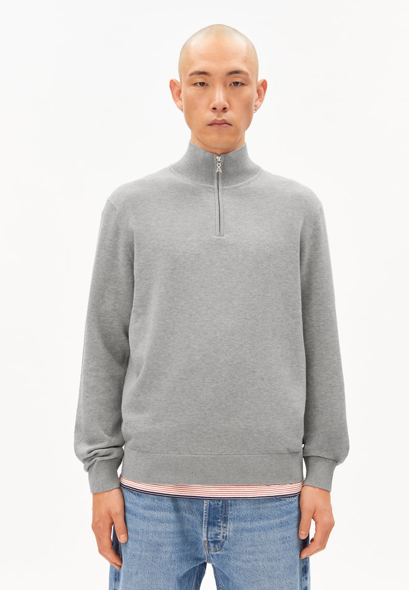 Grauer Zip-up-Pullover mit hohem Kragen, gerippten Bündchen und Saum. Verfügt über ein kleines Logo auf der Brust. Wird über einem gestreiften Hemd und Jeans getragen.