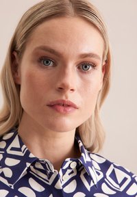 Blusa blu e bianca con motivo geometrico, colletto bottonato e maniche corte. Il tessuto appare liscio con una lieve lucentezza.