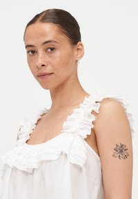 Hvit ermeløs topp med volangstropper, myk tekstur og en avslappet passform. En blomster-tatovering er synlig på modellens overarm.