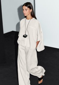 Une personne marche dans une tenue élégante de couleur claire, avec un haut fluide et un pantalon large, accessoirisé d'un collier en pendentif noir.