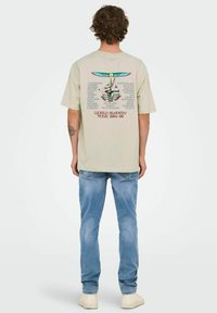 Camiseta de algodón beige con mangas cortas, que presenta un gráfico multicolor en la espalda, que incluye texto y un elemento de diseño estilizado.