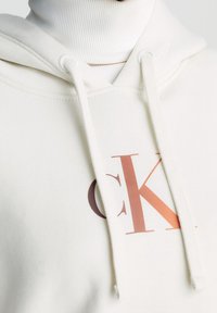 Felpa leggera color crema con cappuccio regolabile, caratterizzata da un grande logo "CK" in rame. Tessuto morbido con un dettaglio di tasca a marsupio.