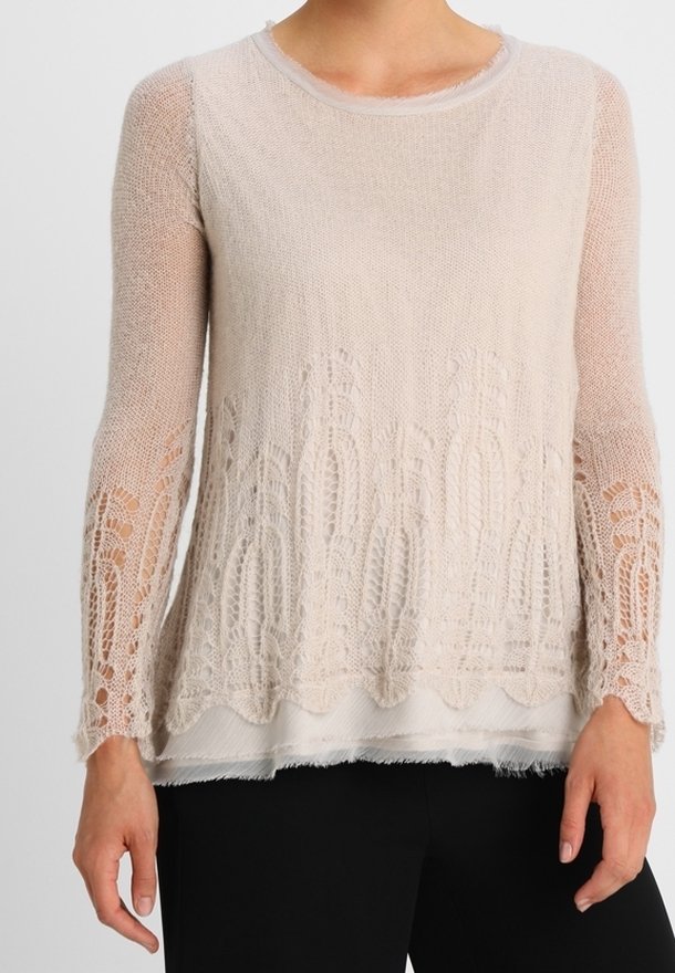 Ljusbeige stickad tröja med långa ärmar, prydd med ett spetsmönster i nederkant och på ärmarna, lager på lager över en vit bas.