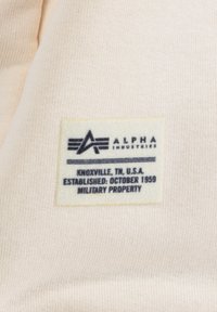 Etiqueta con fondo crema y texto negro, que presenta "ALPHA INDUSTRIES", detalles de ubicación y la frase "PROPIEDAD MILITAR".