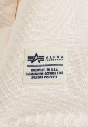 Étiquette avec un fond crème et un texte noir, comportant "ALPHA INDUSTRIES", des détails de localisation et la phrase "PROPRIÉTÉ MILITAIRE".