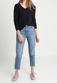 Schwarzer, gerippter Strickpullover mit V-Ausschnitt, kombiniert mit hellblauen Jeans mit hochgekrempelten Bündchen und hellrosa Sneakern. Schlichtes und lässiges Outfit.
