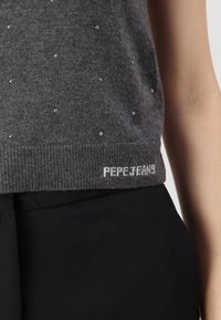 Grå stickad tröja med silverfärgade pärldekorationer, ribbad nederkant och vit "PEPE JEANS"-logotyp nertill, i kombination med svarta byxor.
