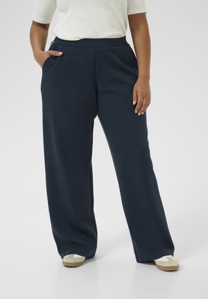 Osoba nosząca granatowe szerokie spodnie, biały top i białe trampki z beżowymi podeszwami, stojąca z jedną ręką w kieszeni.