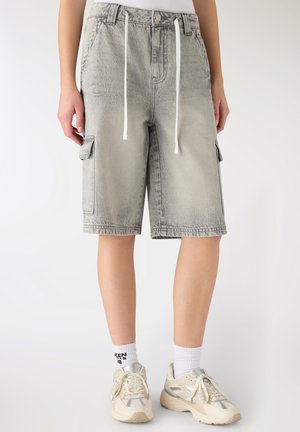 Pantaloncini cargo in denim grigio chiaro con coulisse bianche indossati con calzini bianchi e sneakers beige su una persona in piedi.