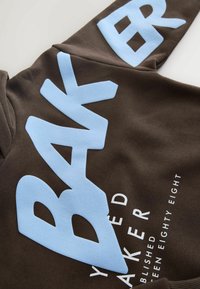Brun sweatshirt med stor lyseblå tekst "BAKER" og hvide tekstdetaljer, med en glat, blød stoftekstur og afslappet pasform.
