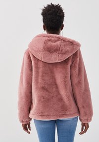 Person iført en pink, fuzzy hættejakke og blå jeans, stående med ryggen mod kameraet foran en ensfarvet lys baggrund.