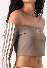 Top cropped castanho com decote à princesa, apresentando três riscas brancas em cada manga e um pequeno logótipo branco na frente. Textura suave.