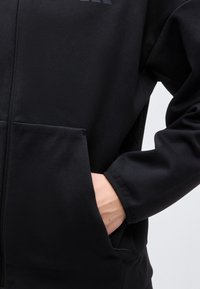 Personne portant une veste noire zippée avec la main partiellement dans une grande poche avant du côté droit.