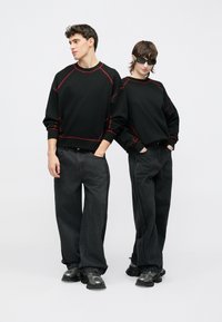 Fekete pulóverek piros varrással, sötét, széles szárú farmerral párosítva. Mindketten vastag fekete cipőket viselnek, és egy egyszerű háttér előtt pózolnak együtt.