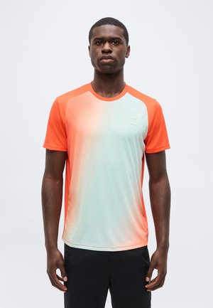 Mladić s kratkom kosom koji nosi kratkih rukava, atletsku majicu u narančasto-svijetloplavom gradijentu i crne hlače, stoji na jednostavnoj pozadini.