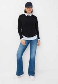 Black sweater over a light blue collared shirt, paired with light blue flared jeans and white sneakers, finished with a navy cap.