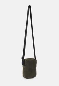 Pieni suorakulmainen oliivinvihreä corduroykäsilaukku, jossa on säädettävä musta hihna, roikkuu yksinkertaista valkoista taustaa vasten.