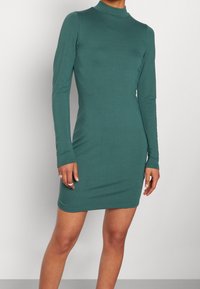 Dopasowana długa zielona sukienka z wysokim dekoltem noszona przez osobę, widoczna od ramion do połowy uda na jednolitym tle.
