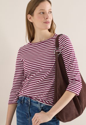 Femme portant un haut à rayures violettes et blanches et un jean bleu, avec un grand sac à bandoulière marron, regardant vers sa gauche.