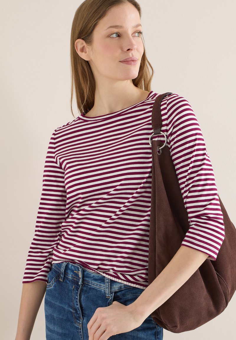 Femme portant un haut à rayures violettes et blanches et un jean bleu, avec un grand sac à bandoulière marron, regardant vers sa gauche.