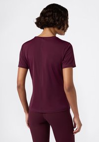 Mujer con cabello rizado corto que lleva una camiseta deportiva ajustada de manga corta en un tono púrpura oscuro y leggings a juego, mostrada de espaldas.