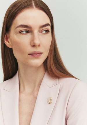 Jonge vrouw met lang bruin haar draagt een lichtroze blazer versierd met een gouden bloemvormige broche, kijkt zijwaarts tegen een neutrale achtergrond.