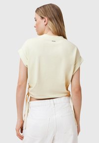 Lichtgele cropped t-shirt van zacht materiaal, met opgerolde mouwen en verstelbare touwtjes aan de zijkanten. Gecombineerd met witte broeken.
