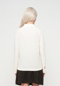 Una persona con largo cabello rubio claro lleva una blusa de mangas largas color crema y una falda marrón oscuro, de pie contra un fondo claro y liso.