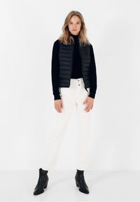 Sort puffervest i lag med en sort sweater, parret med hvide straight-leg jeans og sorte ankelstøvler. En simpel, moderne design.