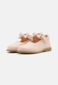 Un par de zapatos para toddlers de color rosa claro con bordes festoneados y correas con moño, suelas beige y cierres de velcro sobre fondo blanco.