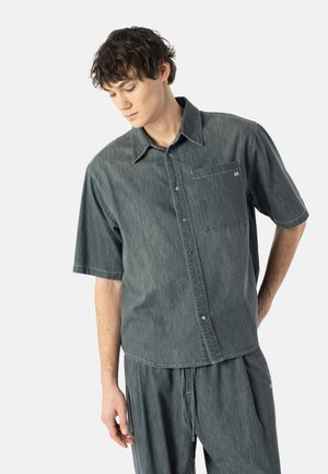 Junger Mann trägt ein locker sitzendes, dunkelgraues Denim-Kurzarmhemd mit Brusttasche und passende Hose, blickt nach unten und hat eine Hand am Rücken.