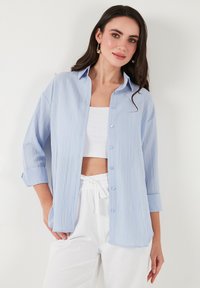 Light blue button-up shirt with a textured design, long sleeves rolled at the cuffs, accompanied by a white crop top and white pants.