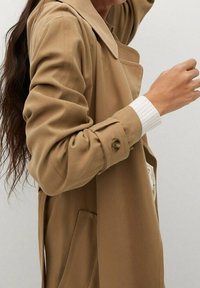 Beige Trenchcoat mit großem Kragen, langen Ärmeln und Knopfakzenten. Weicher, glatter Stoff mit seitlichen Taschen und lässiger Passform.
