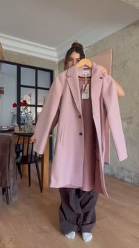 Žena drží na ramienku ružový dlhý kabát v obývacej izbe s drevenou podlahou, jedálenským stolom a červenými kvetmi vo váze za ňou.